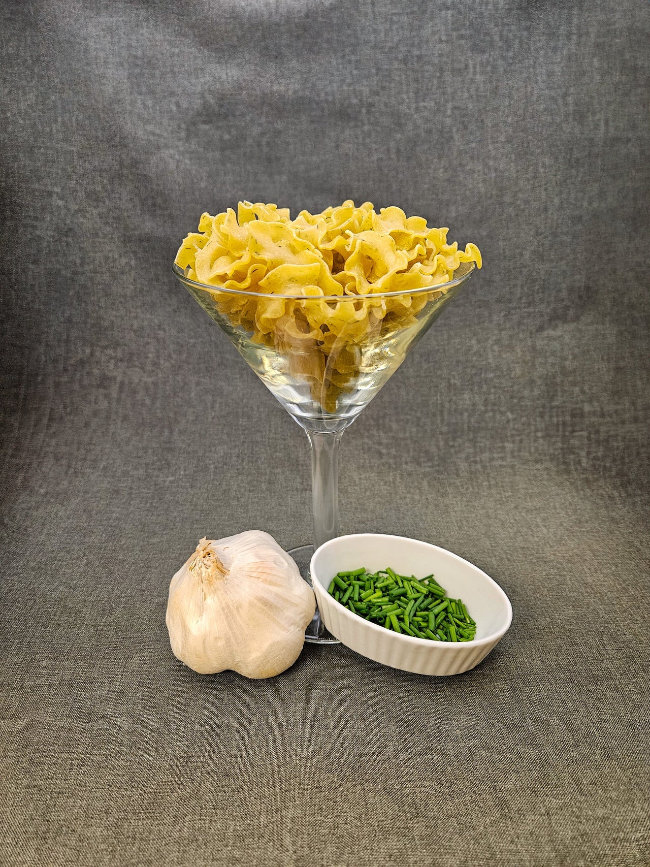 Glass bowl filled with yellow pasta, garlic bulb, and green leafy vegetables on a gray background