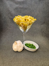 Glass bowl filled with yellow pasta, garlic bulb, and green leafy vegetables on a gray background