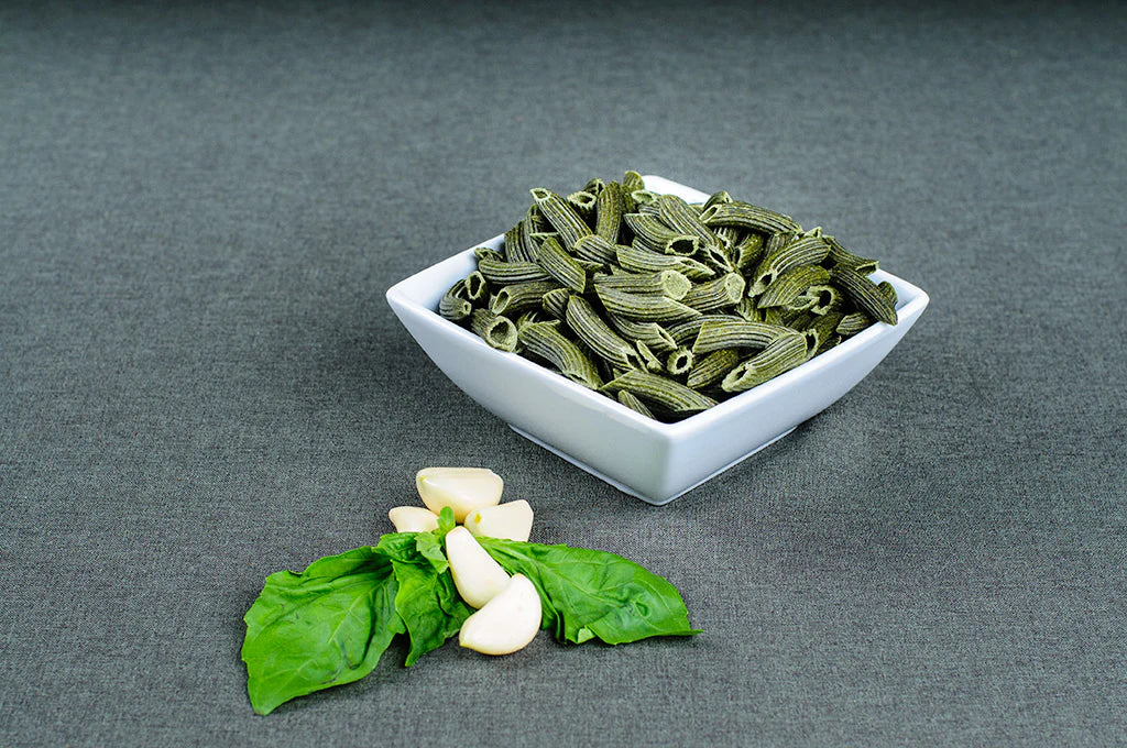 Green pasta in a white bowl on a gray background with basil and garlic.