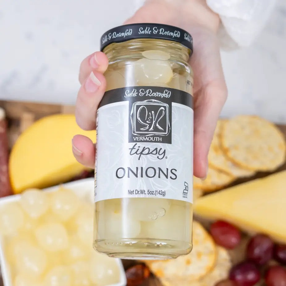 Hand holding a jar of 'Tipsy Onions' vermouth with a platter of cheese and crackers in the background.